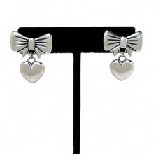 Pair of silver bow and heart earrings on a black earring stand.